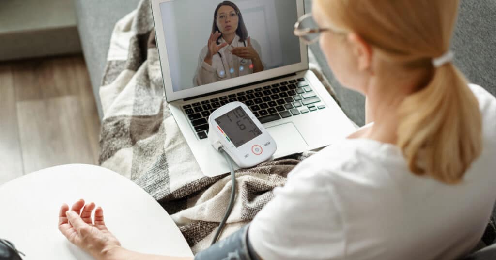 A woman at home using a blood pressure monitor while having a video consultation with a healthcare provider on her laptop. The scene illustrates the concept of Virtual Medicine, highlighting the use of technology to facilitate remote health monitoring and real-time consultations.