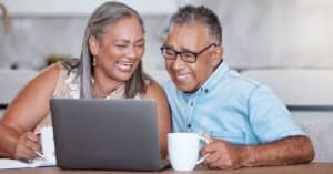 An older couple smiling and looking at a laptop screen while enjoying their coffee at the kitchen table, illustrating the ease and connectivity provided by using a patient portal.