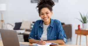 A young woman with curly hair tied up, wearing a blue denim shirt over a white t-shirt, sits at a desk smiling warmly at the camera. She is writing in a notebook with a pen while working on her laptop, illustrating the concept of healthcare navigation and in a cozy, well-lit room with a sofa and plants in the background.