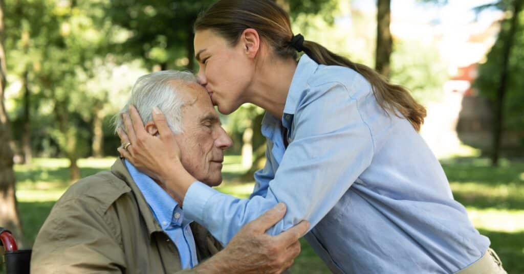 A woman tenderly kisses the forehead of an elderly man sitting outdoors, symbolizing the compassion and support involved in home care.