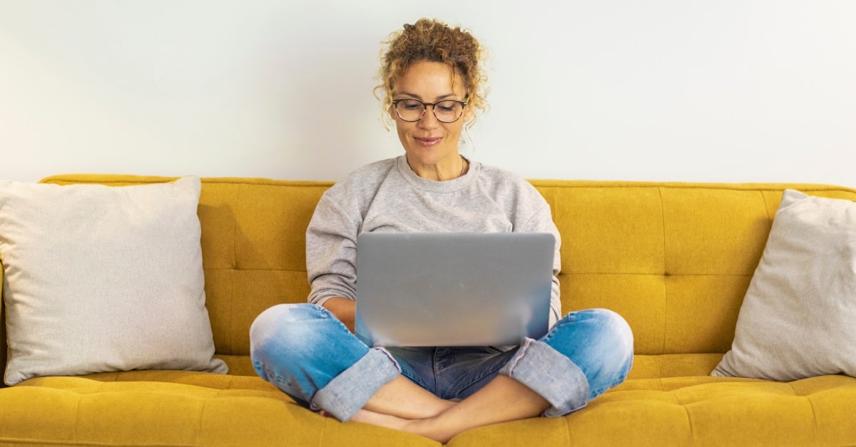 Person sitting cross-legged on a mustard yellow couch, wearing glasses, a gray sweatshirt, and blue jeans, focused on working on a laptop. The setting is cozy, with neutral-colored pillows on either side, creating a comfortable and relaxed environment.