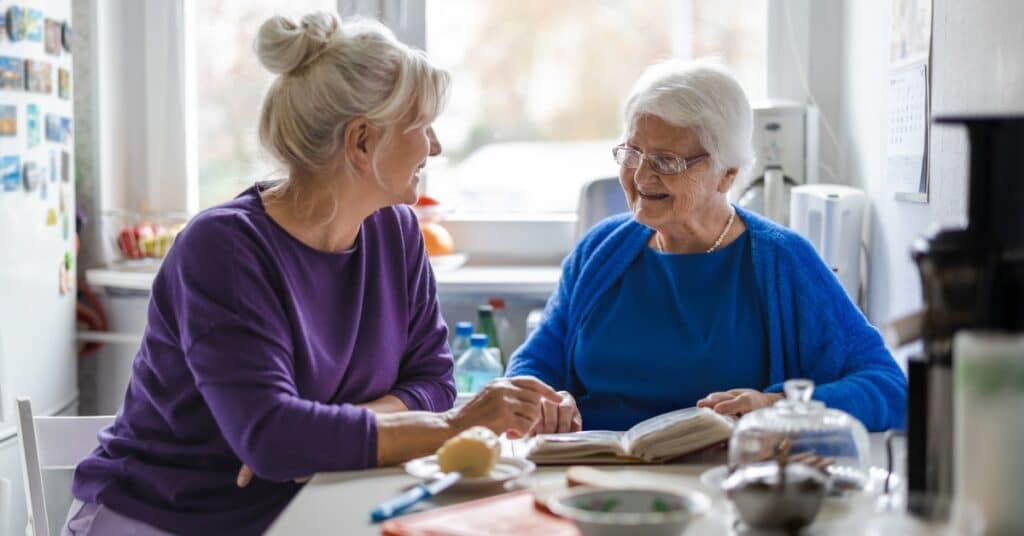 An elderly woman and her caregiver share a warm and engaging conversation at the kitchen table, surrounded by homey details like a cookbook, fruit, and a teapot. This scene illustrates the importance of creating a supportive and calming environment to help manage stress and anxiety in elderly individuals after a health episode.