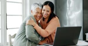 An elderly woman and her adult daughter share a heartfelt embrace while sitting at a kitchen table with a laptop open in front of them, symbolizing connection and mutual support. This image represents the importance of budgeting and forecasting healthcare costs after a diagnosis, as families work together to plan financially for treatments and lifestyle changes. The bright and cozy environment highlights a sense of optimism and collaboration in managing care effectively.