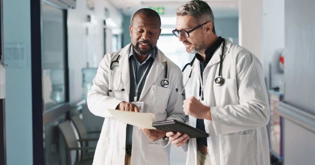 Two healthcare professionals in white coats stand in a hospital hallway, engaged in a focused discussion while reviewing a patient's chart and tablet. Their collaborative posture and attentive expressions highlight the importance of care coordination in ensuring seamless communication and teamwork among providers. The hospital setting reinforces the context of organizing and managing patient care to deliver effective and unified healthcare services.