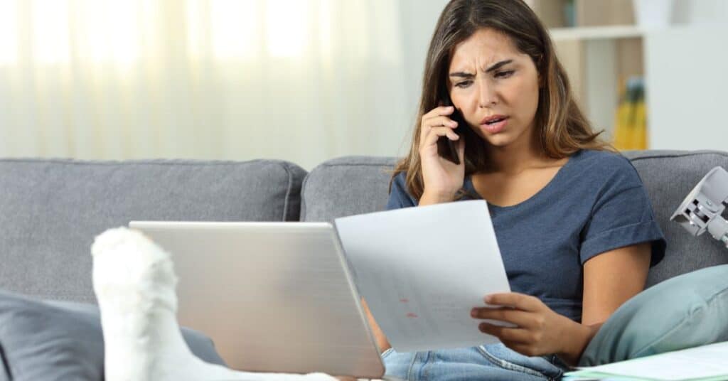 A concerned woman with her leg in a cast sits on a couch, reviewing a medical bill while on the phone, with a laptop open in front of her, illustrating the stress and challenges associated with coverage gaps in healthcare.