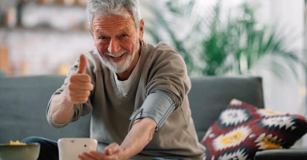 A smiling elderly man sits comfortably on a couch, giving a thumbs up while using a blood pressure monitor, symbolizing confidence and proactive health management. The scene highlights the importance of health literacy, as it empowers individuals to independently monitor their health and make informed decisions. A vibrant, well-lit living room setting emphasizes a positive and supportive environment for self-care.