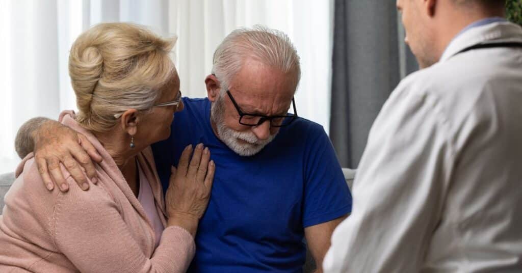 An elderly man receiving a difficult diagnosis sits with emotional support from his companion, while a doctor provides professional guidance in a supportive clinical setting.