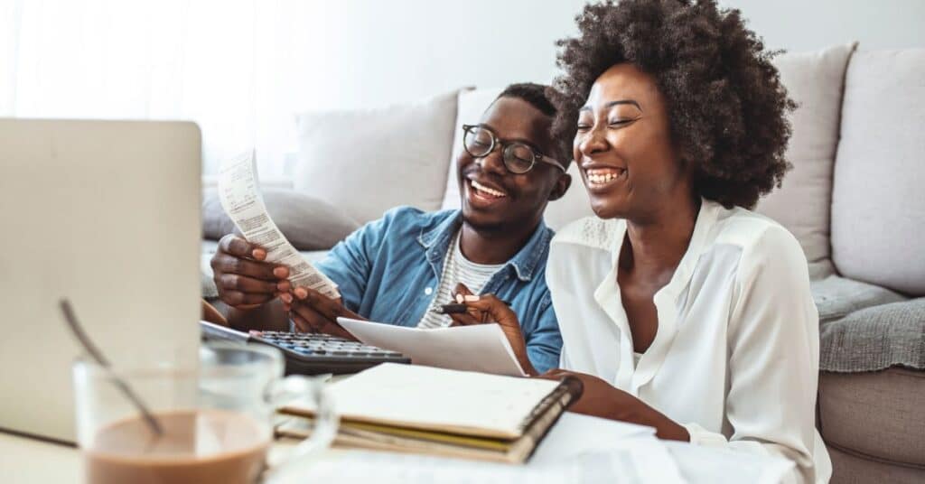 Smiling couple sitting together at home, reviewing medical documents and notes in front of a laptop, symbolizing the process of improving health literacy measurement through self-guided learning. The scene emphasizes patient engagement in healthcare management and the use of online tools to enhance comprehension and navigation of medical information.