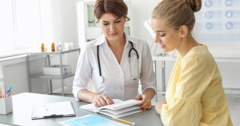 A female doctor in a white coat with a stethoscope discusses treatment options with a smiling female patient in a yellow blouse, both reviewing an open medical document. The setting is a well-lit, modern medical office with a clipboard, pen, and health-related charts in the background. This image represents shared decision-making, where healthcare providers and patients collaborate to make informed choices about care.