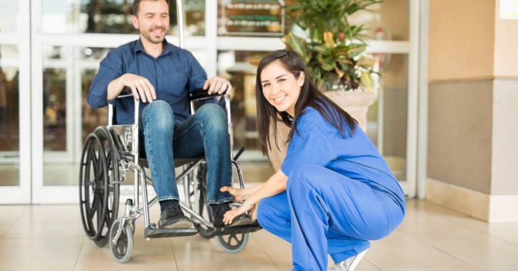 A cheerful female healthcare worker in blue scrubs crouches beside a man in a wheelchair outside a healthcare facility. She is adjusting the wheelchair's footrests with care and smiling warmly at the camera. The man, dressed casually in jeans and a navy button-up shirt, also smiles, appearing at ease and comfortable. Behind them, glass doors and potted plants indicate they are near a hospital or clinic entrance. This compassionate moment illustrates Care Transition: the process of moving a patient from one healthcare setting to another, highlighting the human connection and attentiveness essential to supporting patients during vulnerable times.