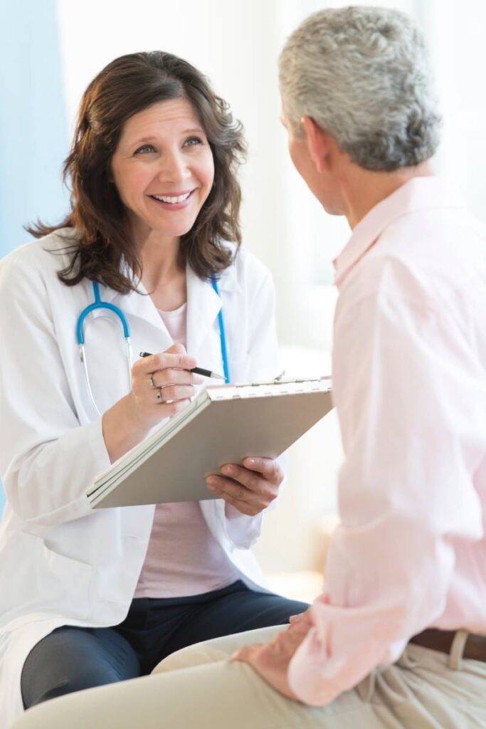 A compassionate female physician in a white coat, with a stethoscope draped around her neck, engages warmly with a middle-aged male patient. She holds a clipboard and pen, attentively listening and smiling as they discuss his health journey. The bright, welcoming medical setting reflects a structured approach to DME patient engagement and compliance, ensuring patients stay informed and actively participate in their care. This image captures the essence of patient-centered support, reinforcing how durable medical equipment providers can enhance compliance, improve outcomes, and streamline healthcare coordination.