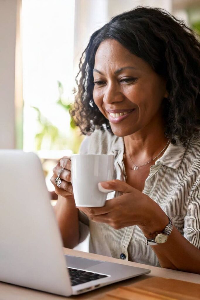 A smiling woman with curly hair is sitting at a table, holding a white coffee mug in one hand while engaging with her laptop. She appears relaxed and content, representing an engaged health plan member who is actively managing her healthcare. The bright, natural lighting and comfortable home setting reflect how health insurers prioritize member satisfaction and engagement by providing seamless digital tools, proactive outreach, and self-guided health literacy programs. The image conveys the idea that well-informed members make better healthcare decisions, leading to improved compliance, reduced costs, and stronger retention for insurers.