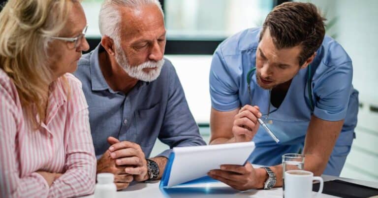 A middle-aged male healthcare provider in blue scrubs is sitting at a table with an older couple, attentively explaining medical information using a clipboard and pen. The elderly man and woman, appearing thoughtful and engaged, listen closely as they review documents, with a bottle of medication and glass of water on the table. This scene visually represents the concept of “how do I prepare questions to ask my doctor” by illustrating a collaborative and informed conversation during a medical consultation.
