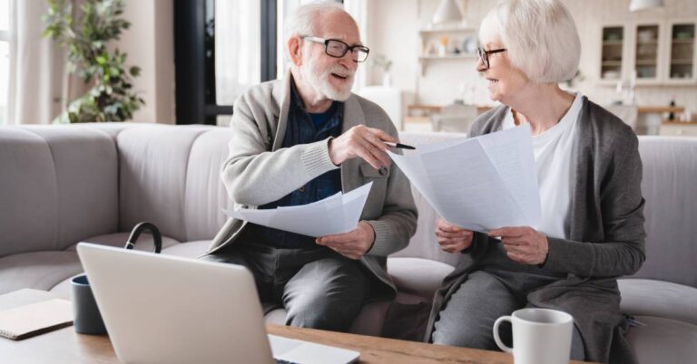 An older couple sits on a light-colored sofa in a cozy living room, reviewing paperwork together with a laptop open in front of them. The man, smiling, gestures toward a document while the woman looks at him attentively, holding another page. A coffee mug, notepad, and glasses rest on the table, suggesting a thoughtful planning session. The image illustrates proactive health management, reflecting the question: What steps can I take to avoid medical oversights? It conveys how preparation, shared decision-making, and staying organized can help prevent costly or dangerous errors in medical care.