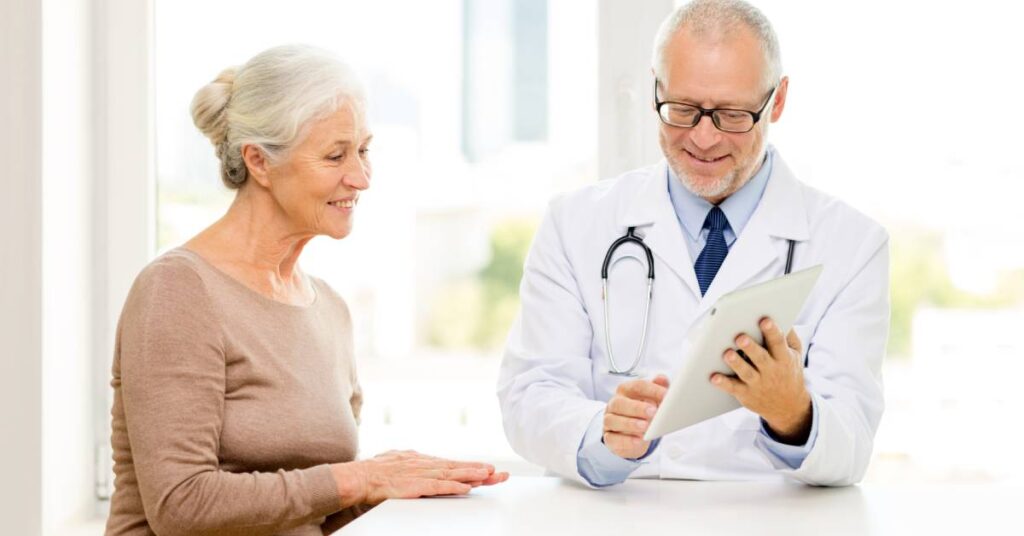 An older woman in a tan sweater sits at a bright desk in a sunlit exam room, her gray hair pulled back as she smiles and rests her hands on the tabletop; across from her, a male doctor in a white coat and tie holds a tablet and gestures to review her chart—this scene depicts a follow-up appointment, a scheduled visit after your initial consultation or treatment to monitor your progress, review any test results, adjust your care plan as needed, and address any new questions.