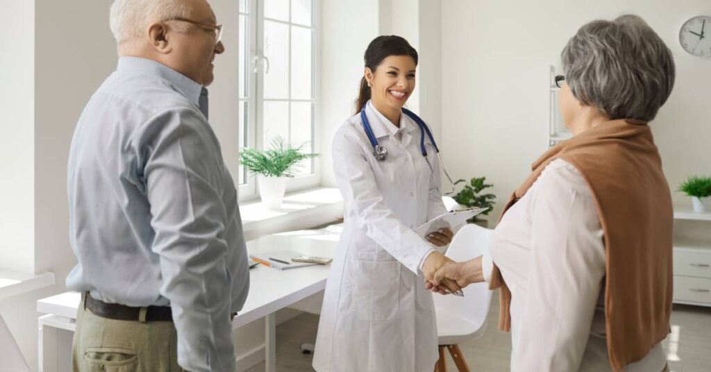 An elderly woman shakes hands with a smiling female doctor in a bright exam room—with the woman’s husband standing nearby, a clipboard, stethoscope, houseplant on the windowsill, and wall clock visible—illustrating a medical appointment, a scheduled visit with a healthcare provider where you receive evaluation, diagnosis, or treatment without being admitted to the hospital.