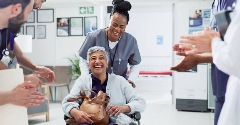A joyful older woman with short gray hair sits in a wheelchair, beaming with a wide smile as a nurse in scrubs stands behind her, pushing the chair down a hospital hallway. The nurse is also smiling, and both are surrounded by clapping healthcare professionals in white coats and stethoscopes, forming a cheerful send-off. The woman clutches a tan handbag on her lap, radiating confidence and happiness. This image powerfully captures Patient Satisfaction — the measure of how well a patient's healthcare experience met their needs, expectations, and preferences — reflected in the patient's emotional relief and the supportive energy of her care team.