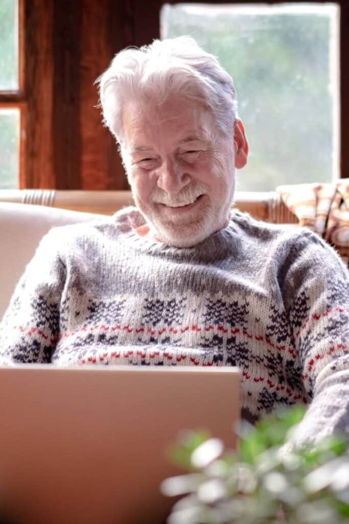 An older adult sits comfortably on a couch in a cozy, sunlit room, smiling warmly while engaging with a laptop. This image represents a patient who has been empowered to take control of their health through education—not through tech gimmicks. The photo symbolizes how CMS Star Ratings improve when members are taught to actively participate in their care. Unlike passive tools like advanced patient portals, frequent reminders, or impersonal surveys, this moment illustrates the power of behavior change—showing that meaningful engagement comes from learning how to navigate healthcare, not just clicking through it.