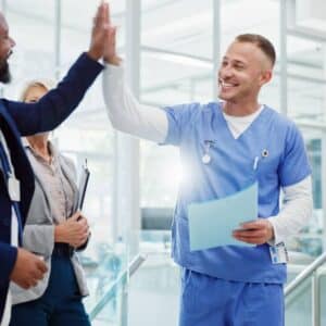 A healthcare administrator in a business suit enthusiastically high-fives a smiling male nurse in blue scrubs, while a female nurse looks on with approval. The scene takes place in a modern, sunlit hospital or clinical setting. The mood is upbeat and collaborative, representing a successful implementation of the PatientBetter Licensing Suite Start-up Kit—an engagement and compliance solution designed for healthcare professionals to elevate patient outcomes through teamwork and strategic support.