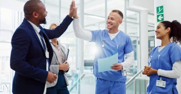 A healthcare administrator in a business suit enthusiastically high-fives a smiling male nurse in blue scrubs, while a female nurse looks on with approval. The scene takes place in a modern, sunlit hospital or clinical setting. The mood is upbeat and collaborative, representing a successful implementation of the PatientBetter Licensing Suite Start-up Kit—an engagement and compliance solution designed for healthcare professionals to elevate patient outcomes through teamwork and strategic support.