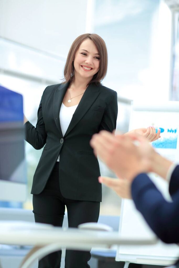 A confident female healthcare administrator stands in a modern conference room, smiling as she receives applause from colleagues. She is dressed professionally in a dark blazer and stands in front of a flip chart displaying upward-trending data. The setting conveys strategic success and leadership. This image reflects the empowered role of healthcare administrators in driving modern MIPS compliance and quality assurance—highlighting Patient Better’s licensing solution as a smart, measurable, and leadership-driven approach to post-EHR patient engagement and reporting.