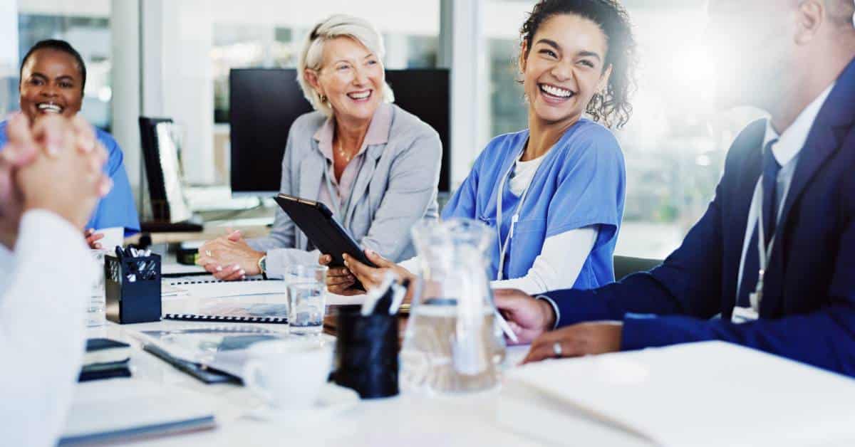 A diverse group of healthcare and administrative professionals sits around a conference table in a bright, modern office. A smiling nurse in scrubs engages confidently with her colleagues, including an older woman in business attire holding a tablet and a man in a suit. Laptops, documents, and water glasses are spread across the table, suggesting a collaborative planning session. The group exudes optimism, trust, and forward-thinking strategy. This scene reflects the value of Patient Better’s licensing tool—where modern, post-EHR MIPS compliance is no longer a burden, but a smart, patient-led solution. By streamlining quality reporting and documentation, Patient Better empowers administrators to meet regulatory standards efficiently while enhancing care coordination and staff confidence.