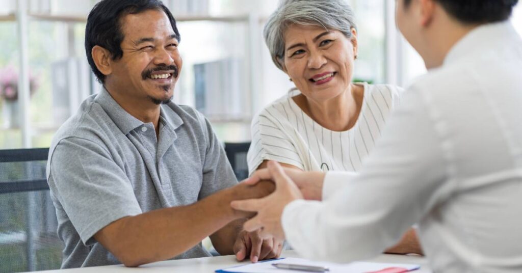 A smiling older man and woman sit across a desk from a healthcare provider in a bright, welcoming medical office. The man, wearing a light gray polo shirt, is shaking hands confidently with the provider, who is dressed in a white coat. The woman, in a white pinstriped blouse, looks on warmly, symbolizing trust and satisfaction in the care being received. This image represents a positive Patient Experience — the full range of interactions, perceptions, and emotional responses a patient has while navigating the healthcare system, including communication, comfort, and care outcomes.