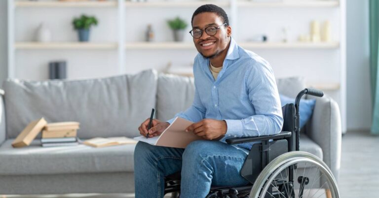 A smiling man in a wheelchair sits at a table in a cozy living room, wearing glasses and a light blue shirt, holding a pen while writing in a notebook. Sunlight filters into the space, and books and plants decorate the shelves behind him, creating a warm, comfortable atmosphere. This image represents quality of life—the overall sense of well-being, including physical health, emotional balance, social connection, and the ability to engage in meaningful daily activities.