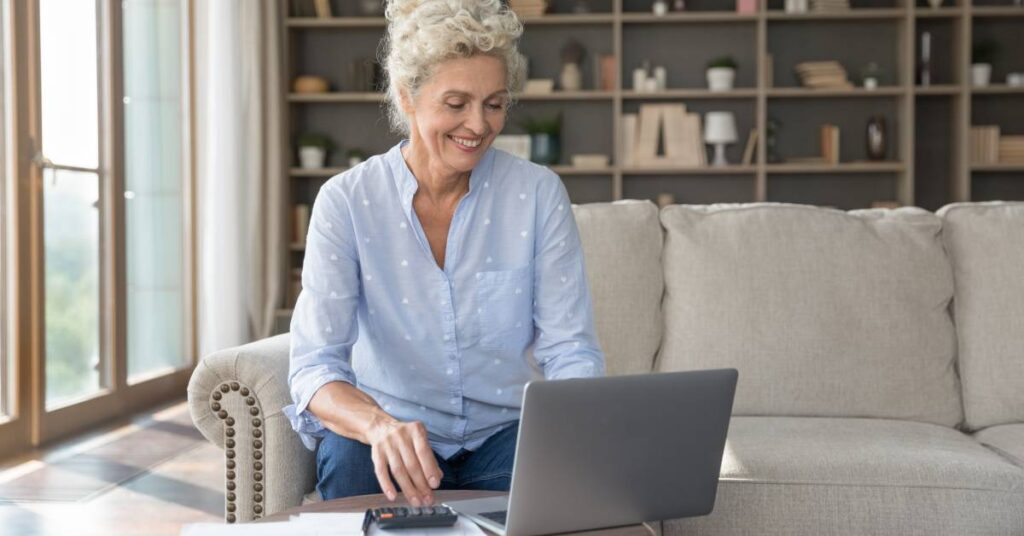 Smiling older woman reviewing her explanation of benefits on a laptop at home, sitting on a comfortable couch with paperwork and a calculator nearby, symbolizing confident and independent healthcare management.