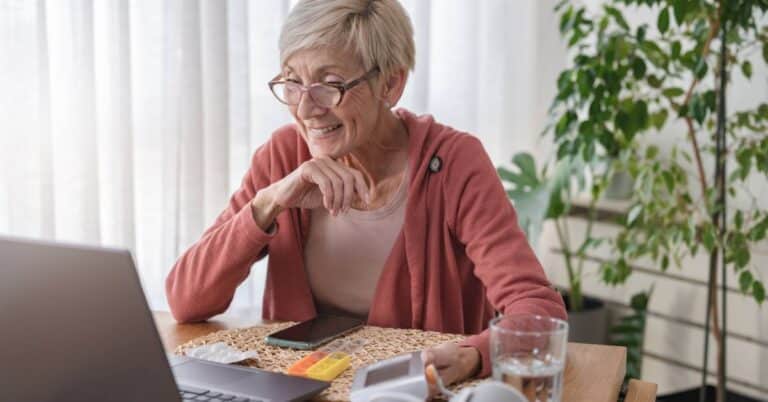 An older woman with short gray hair and glasses, wearing a rose-colored cardigan, sits at a kitchen table smiling during a video call on her laptop. In front of her are a pill organizer, a blood pressure cuff, a smartphone, and a glass of water—suggesting she is discussing her health or medications online. The scene illustrates a patient engaging in virtual care, possibly learning about or seeking preauthorization for medical services or prescriptions.