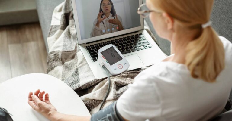 A woman uses a blood pressure monitor at home while speaking with a healthcare provider via video call on her laptop, illustrating Digital Therapeutics in a virtual care setting.