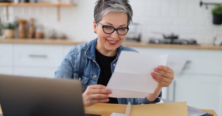 A smiling woman sits at her kitchen table using a laptop and reading a letter, representing the convenience and accessibility of virtual-first care from home.