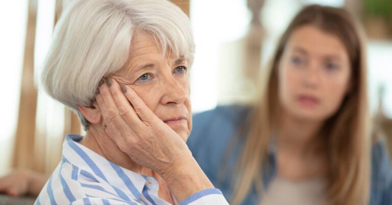 Burden of care shown by a tired older woman with caregiver in background.