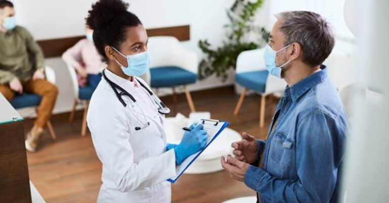 A doctor speaks with a patient in a waiting room while others wait in the background, illustrating risk stratification as providers assess and prioritize care needs.