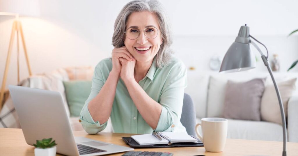 Older woman sitting at a desk with a notebook, smiling confidently as she prepares her signpost questions for an upcoming medical appointment.