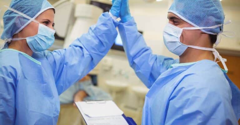 Two critical care specialists in surgical gowns and masks high-five in a hospital setting, celebrating teamwork and successful patient care.