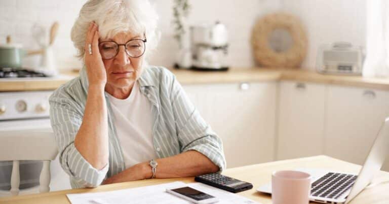 Older woman reviewing her medical bill at home with concern, calculator and laptop on the table.