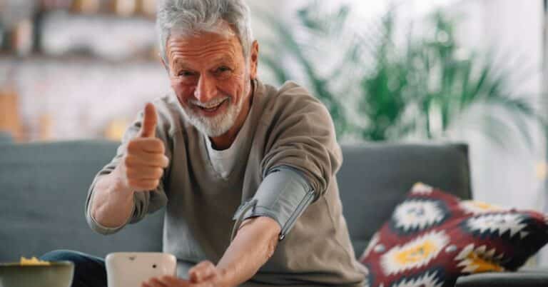 An older man smiling and giving a thumbs up while checking his blood pressure at home, representing cardiology and heart health management.