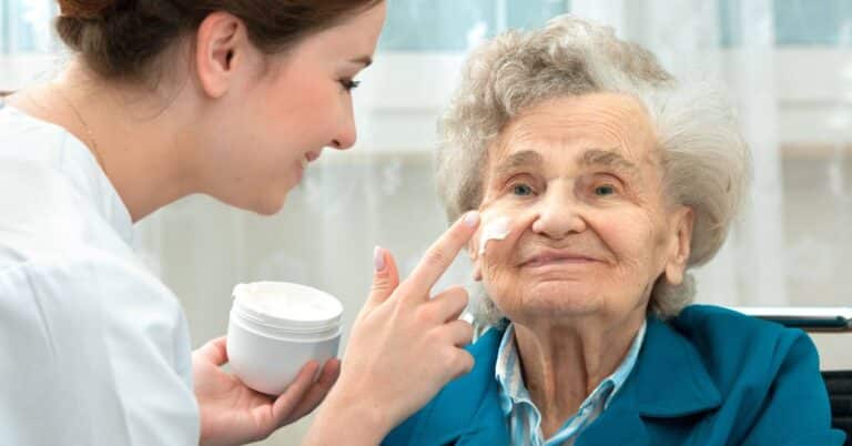 A healthcare professional applying a moisturizing cream to an older woman’s face, representing dermatology care, skin health, and compassionate skin treatment.