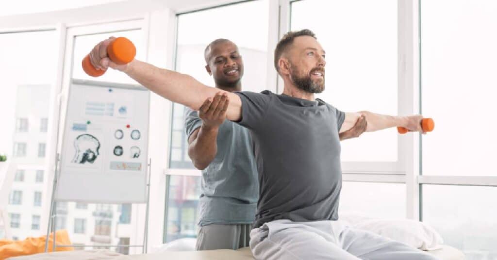 A physical therapist assisting a middle-aged man with arm exercises using small weights during a rehabilitation session, demonstrating physical therapy for strength, balance, and mobility recovery.