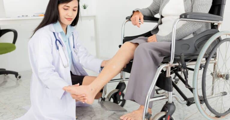 A podiatry specialist examines an elderly patient’s leg and foot while the patient sits in a wheelchair, illustrating compassionate podiatric care and mobility support in a clinical setting.