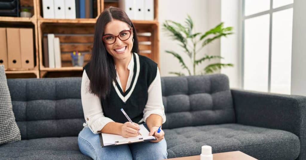 A smiling psychologist sitting on a couch with a clipboard, representing psychology, therapy, and mental health counseling in a supportive setting.