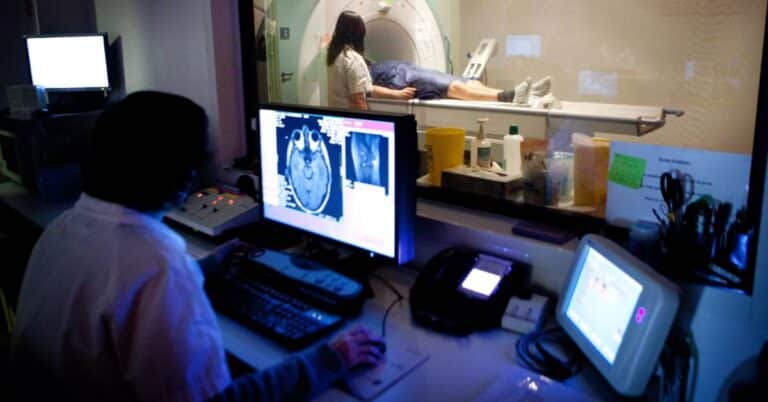 A radiology technician monitors brain scan images on a computer while a patient undergoes an MRI, illustrating how radiology uses imaging to diagnose medical conditions.