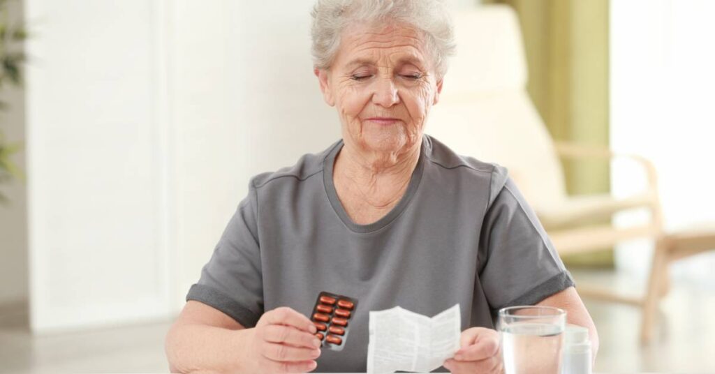 An older woman reads a medication package insert at home, reviewing the warning label before taking her prescribed pills.