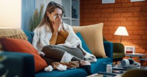 A woman sitting on a couch with tissues and cold supplies, appearing sick as she manages symptoms related to the New Flu Strain 2025.