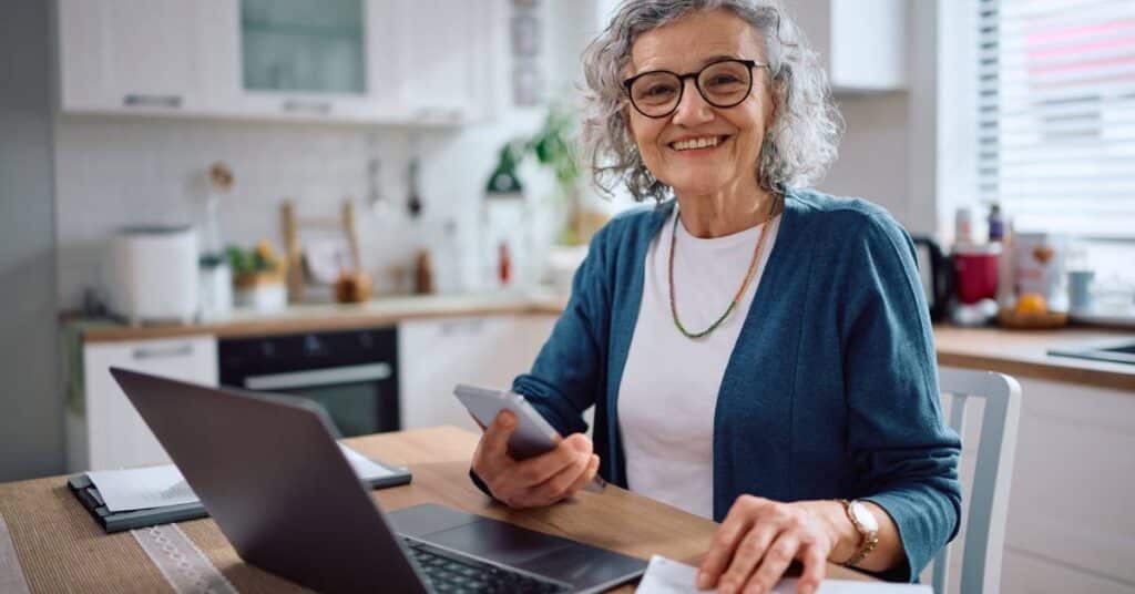 Smiling older woman using her laptop and phone at home to manage her healthcare, illustrating the Patient Better vs WebMD difference by showing how real navigation and care coordination skills support today’s medical needs.