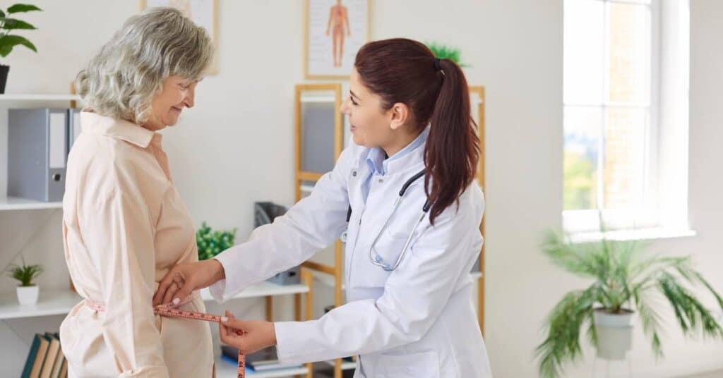 Doctor measuring an older patient’s waist during a consultation about the Trump drug pricing deal and new Medicare coverage for weight-loss medications.