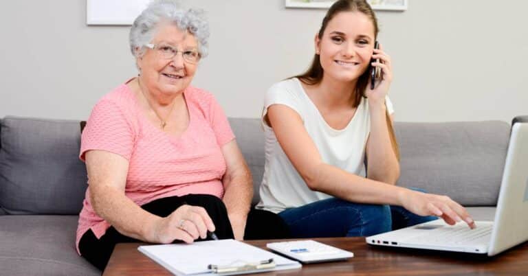 Caregiver helping an older adult review papers and update clinical documentation at home using a laptop and phone.