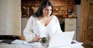 A woman sitting at her kitchen table with a laptop and notes, practicing how to track symptoms by organizing her health information at home.