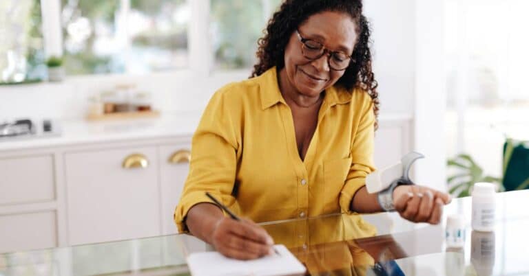 A woman reviews her medications at home and writes an organized medication list while checking her blood pressure.